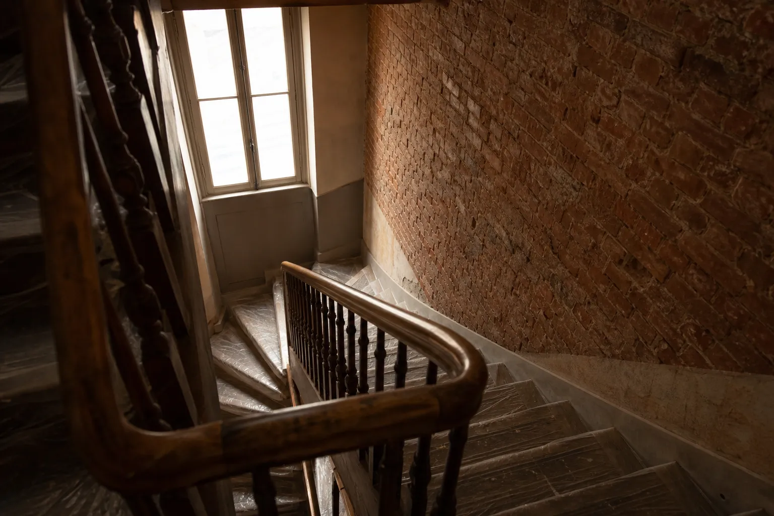 Cage d’escalier d’un immeuble ancien, marches en bois protégées par un film de chantier, rampe en bois, mur en brique, lumière d’ouest par u